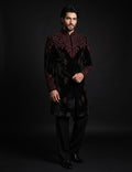 A man modeling a black velvet sherwani jacket with intricate dark red embroidery, paired with black trousers and black dress shoes, set against a dark studio background.