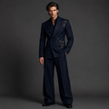 A man modeling a dark navy double-breasted suit with ornate gold embroidery on the lapels and shoulders, paired with matching trousers, a black shirt, and black dress shoes against a dark gray studio background.