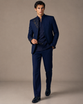 A man modeling a dark navy blue three-piece suit with gold embroidery on the lapel and sleeve, paired with a matching navy shirt and trousers, against a solid beige studio background.