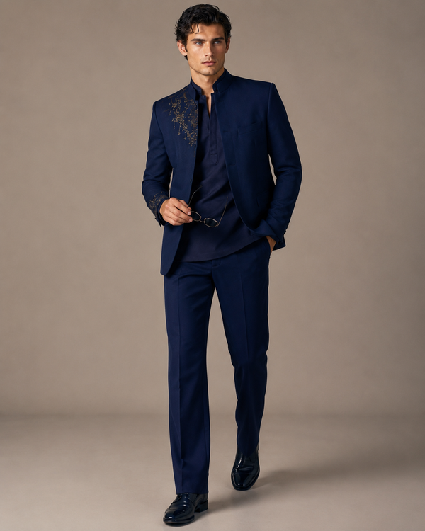 A man modeling a dark navy blue three-piece suit with gold embroidery on the lapel and sleeve, paired with a matching navy shirt and trousers, against a solid beige studio background.
